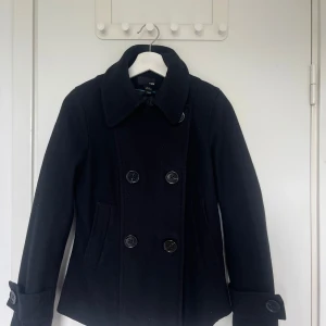 Svart skepparkavaj från H&M - Klassisk svart skepparkavaj från H&M med dubbelknäppning och stora knappar framtill. Jackan har bred krage, två snedställda fickor och dekorativa knappar vid ärmsluten. Perfekt för dig som gillar en stilren och tidlös look.