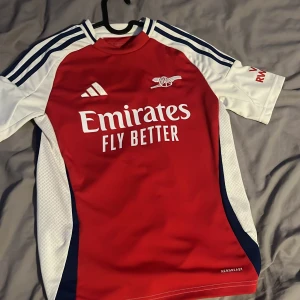 Arsenal Adidas fotbollströja röd/vit - Snygg Arsenal fotbollströja från Adidas i rött och vitt med blå detaljer. Klassisk design med klubbmärke och Emirates Fly Better-tryck på bröstet. Kortärmad modell i lätt och ventilerande material, perfekt för träning eller match. Aldrig använd