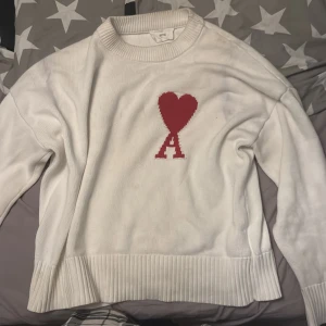 Vit stickad tröja med rött hjärta från Ami - Vit stickad tröja från Ami med ett stort rött hjärta och bokstaven A på bröstet. Tröjan har ribbade muddar och rund hals. Perfekt för dig som gillar stilrena plagg med en twist priset kan diskuteras 🤩
