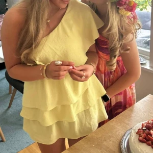 Gul omlottklänning med volanger - Superfin ljusgul kortklänning med volanger och oneshoulder-modell. Klänningen har flera lager och en snygg omlottkänsla som ger extra volym. Perfekt för sommarens alla tillfällen och riktigt skön att bära.