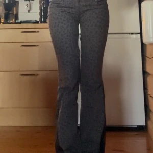 Grå leopardmönstrade bootcut jeans - Säljer ett par grå jeans med leopardmönster och bootcut-snitt. Byxorna har låg midja, klassiska fem fickor och stängs med dragkedja och knapp. Materialet är stretchigt och bekvämt, perfekt för dig som vill sticka ut lite extra.