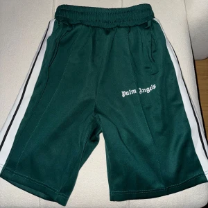 Palm Angels shorts - Snygga gröna shorts från Palm Angels med vita och svarta ränder längs sidorna. De har elastisk midja med snörning och logga broderad på ena benet. Perfekta för en sportig och trendig look.