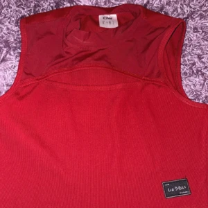 Röd ärmlös linnetopp från KIW - Snygg röd ärmlös linnetopp från iciw med ribbad struktur och en liten svart patch med japansk text framtill. Toppen har en sportig vibe och hög halsringning, perfekt att styla med jeans eller kjol eller tränna .