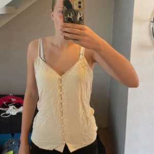 Beige linnetopp med knappar Gina Tricot - Supersöt beige linnetopp från Gina Tricot med smala axelband och dekorativa knappar framtill. Toppen har v-ringning och är figurnära med en lätt avslappnad passform. Perfekt för varma dagar och enkel att styla till olika looks.💛