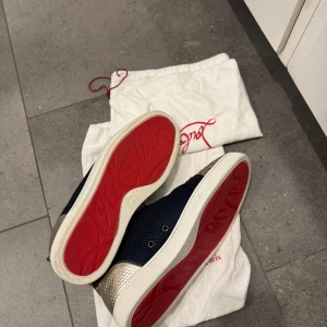 Christian Louboutin sneakers med glitter - Snygga sneakers från Christian Louboutin med klassisk röd sula, blå mesh på sidorna, svarta detaljer och glittrig tå i guldton. Baksidan har metallic guldskinn med struktur. Skorna har snörning och rund tå, perfekta för dig som vill sticka ut. Kartong medföljer ej.