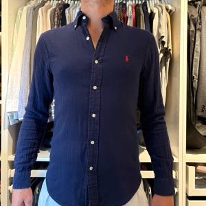 Mörkblå skjorta från Ralph Lauren - Klassisk mörkblå skjorta från Ralph Lauren med lång ärm och knappar framtill. Skjortan har button-down krage och den ikoniska röda logotypen broderad på bröstet. Tillverkad i mjuk bomull för en bekväm känsla och snygg passform.
