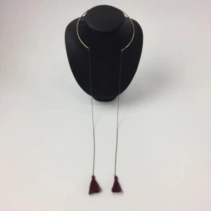 Guldfärgat stelt halsband med vinröda tofsar - Unikt halsband med stel guldfärgad ring som vilar runt halsen och tunna kedjor som avslutas med två vinröda tofsar. Halsbandet har en modern och trendig design som sticker ut.