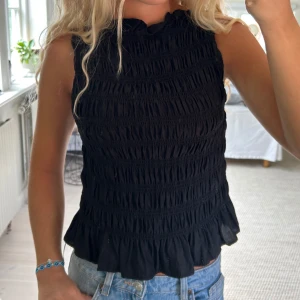 Svart smockad ärmlös topp med volang - Supersöt svart ärmlös topp med smockad struktur och volangkant nertill. Hög halsringning och lätt croppad passform gör den perfekt till jeans eller kjol. Tunn och luftig, passar perfekt för varma dagar.