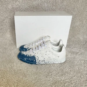 Maison Margiela sneakers  - Maison Margiela sneakers i vitt skinn med unika blå färgstänk över tå och sidor. Klassisk låg modell med snörning och vit sula. Snygg och konstnärlig design som sticker ut, perfekt för dig som gillar statement-plagg.‼️Endast använda en gång ‼️