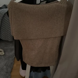 Beige stickad offshoulder tröja Adoore - Supersnygg beige tröja från Adoore med bred ribbad offshoulder-krage. Tröjan är figurnära och har en mjuk, stickad känsla som gör den både trendig och bekväm. Perfekt för dig som gillar en stilren och chic look.