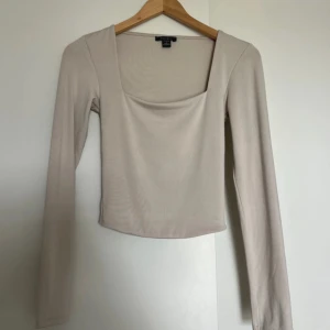 Beige långärmad topp från NewYorker - Säljer en stilren beige långärmad topp från NewYorker med fyrkantig urringning. Toppen är croppad och har en tight passform som sitter snyggt på kroppen. Perfekt att matcha med jeans eller kjol för en clean look.