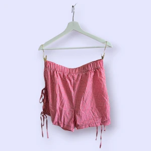 Rödvitrutiga shorts med snörning - Superfina rödvitrutiga shorts med snörning i sidorna som ger en unik och lekfull look. De har elastisk midja och är tillverkade i ett lätt bomullstyg, perfekta för varma dagar. Passformen är loose och mönstret är klassiskt rutigt.