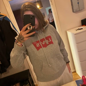 Grå Levi's hoodie med röd logga - Säljer en grå hoodie från Levi's med klassisk röd logga framtill. Tröjan har huva med snörning, magficka och långa ärmar. Något kort i modell och armar 