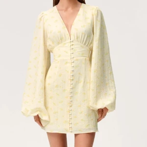 Florence kort blommig klänning - Superfin ljusgul kort klänning med små blommor, v-ringning och markerad midja. Klänningen har långa puffärmar och knappar framtill som ger en söt vintagekänsla. Perfekt för sommarens alla tillfällen!