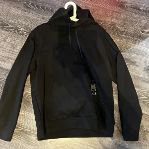 Svart hoodie 1017 ALYX 9SM med spänne - Svart hoodie från 1017 ALYX 9SM med klassisk huva, dragsko och känguruficka. Unik detalj med metallspänne och D-ring på fickan. Raka ärmar och relaxed fit, perfekt för en clean och modern streetwear-look.