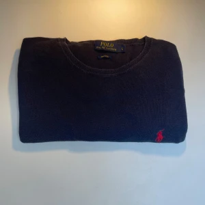 Mörkblå stickad tröja Polo Ralph Lauren - Klassisk mörkblå stickad tröja från Polo Ralph Lauren i mjuk pima-bomull. Tröjan har rund halsringning och ikonisk röd broderad logga på bröstet. Perfekt för dig som gillar stilren och tidlös design.