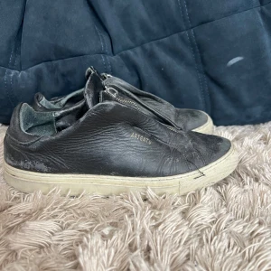 Svarta sneakers från Axel Arigato - Svarta sneakers i skinn från Axel Arigato med vit sula och cool dragkedja framtill istället för klassisk snörning. Minimalistisk design med diskret logga i guld på sidan. Perfekta för dig som gillar stilrena och unika skor.