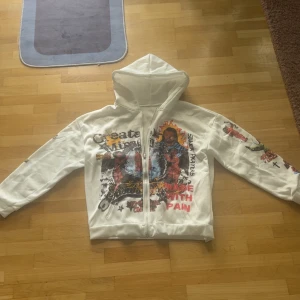 Vit hoodie med tryck och dragkedja - Vit hoodie med färgglada tryck och texter på framsidan och ärmarna. Tröjan har huva, hel dragkedja och ribbade muddar. Snygg streetstil med unika detaljer och motiv som sticker ut. Perfekt för dig som gillar att synas och vill ha en hoodie med attityd.