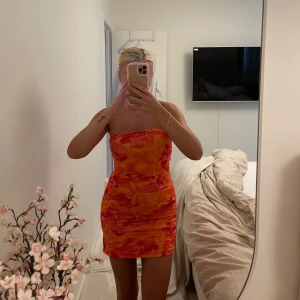 Sommarklänning - Säljer en färgstark tubklänning från SHEIN i orange och rosa toner med blommigt mönster. Klänningen är kort, figurnära och har rynkade detaljer längs sidorna. Perfekt för dig som vill sticka ut med en somrig vibe!