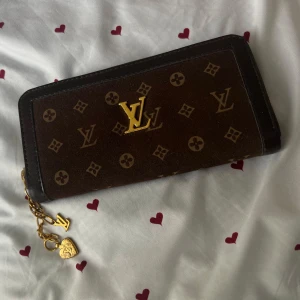 brun plånbok med gulddetaljer - Säljer en brun plånbok med klassiskt monogrammönster och guldfärgade detaljer. Plånboken har dragkedja med snygga berlocker, bland annat ett hjärta och logga. Perfekt storlek för att hålla koll på kort och cash.