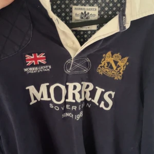 Morris Gents långärmad rugbytröja M - Mörkblå långärmad rugbyinspirerad skjorta från Morris Gents med vit krage, broderad brittisk flagga och guldfärgat emblem på bröstet. Snygga quiltade detaljer på armbågarna och klassisk knappslå framtill. Perfekt för dig som gillar preppy stil.