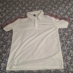 Vit pikétröja med rutiga axlar - Vit pikétröja med klassisk krage och knappar framtill. Axlarna har snygga rutiga detaljer i rött, svart och vitt som ger en unik touch. Tröjan har korta ärmar och en liten broderad logga på bröstet. Perfekt för en clean och stilren look.