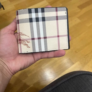 Burberry plånbok - Säljer en snygg plånbok från Burberry i klassiskt rutigt mönster med beige, svart och röda toner. Utsidan har den ikoniska Burberry-rutan och insidan är svart med flera kortfack och fönster för ID. Tillverkad i äkta skinn och känns riktigt lyxig.