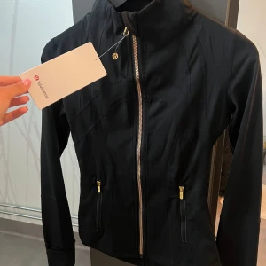 Svart träningsjacka från Lululemon - Svart, figursydd träningsjacka från Lululemon med snygga guldiga dragkedjor och hög krage. Jackan har långa ärmar och två fickor framtill med dragkedja. Perfekt för dig som vill ha en sportig och stilren look.