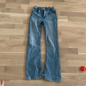 Blå bootcut jeans från Kappahl, stl 140 - Snygga ljusblå jeans från Kappahl i storlek 140. Modellen har bootcut-ben och klassisk femficksdesign. Jeansen är i denim med slitningar och hål på benen för en cool, avslappnad look.