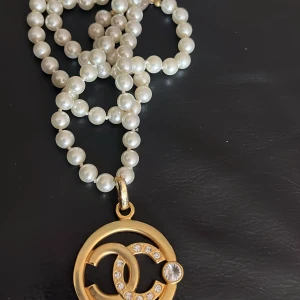 Chanel pärlhalsband med guldplaterad CC-hänge - Lyxigt halsband från Chanel med vita pärlor och ett stort guldigt CC-hänge dekorerat med gnistrande stenar. Hängsmycket har den ikoniska Chanel-loggan och en extra stor kristall. Perfekt statement piece för dig som älskar exklusiva accessoarer.