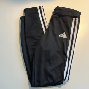 Svarta Adidas träningsbyxor med ränder, barnstorlek. - Svarta träningsbyxor från Adidas med klassiska vita ränder längs sidorna och stretchiga midja. Byxorna har en tight passform med stretchig material som passar för träning. Nötta längs midjan på framsida. *Storleken har suddats ut, men passar för barn runt 140 cm