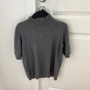 Grå polotröja med kort ärm från H&M - Snygg grå polotröja från H&M med ribbad hög krage och korta ärmar. Tröjan är i mjukt stickat material och har en liten metallring som detalj i nacken. Skriv om ni har frågor! :)