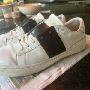 Valentino Garavani Open sneakers vit/svart - Snygga Valentino Garavani Open sneakers i vitt skinn med bred svart läderdetalj över snörningen. Klassisk rund tå, platt sula och diskreta nitar baktill. Perfekta för dig som gillar stilrena och lyxiga sneakers med en twist. Priset går att diskutera👍hör av er om ni har några frågor😊