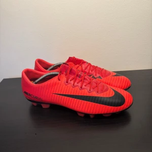 Nike Mercurial röda fotbollsskor storlek 46 - Snygga Nike Mercurial fotbollsskor i rött med svarta detaljer och stora Nike-loggor på sidorna. Skorna har snörning, platt sula med dobbar och är tillverkade i syntetmaterial för bästa grepp och känsla på planen.