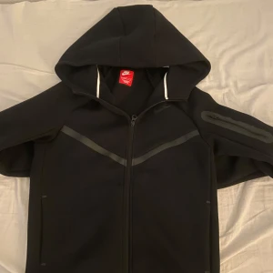 Svart Nike hoodiejacka med dragkedja - Svart hoodiejacka från Nike med hel dragkedja och huva. Jackan har diskreta svarta detaljer, en ficka med dragkedja på ärmen och sportig look. Perfekt för dig som gillar streetwear och vill ha något bekvämt och stilrent.