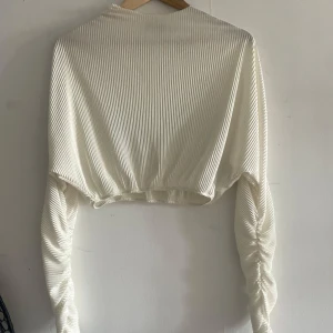 Vit ribbad croppad topp Bianca x Nelly - Säljer en vit croppad topp från Bianca x Nelly med långa ärmar och ribbad struktur. Toppen har en lätt glansig finish och är i ett stretchigt material som sitter snyggt. Perfekt till jeans eller kjol för en trendig look.