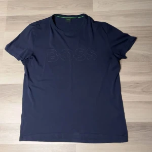 Hugo boss T-shirt - Snygg mörkblå t-shirt från BOSS med diskret BOSS-logga ton-i-ton på bröstet. Klassisk rund halsringning och korta ärmar. Tillverkad i mjuk bomull som känns skön mot huden. Perfekt för en clean och stilren look.