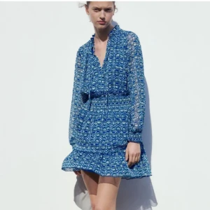 Blå mönstrad kortklänning från Zara - Supersöt blå kortklänning från Zara med vitt och grönt mönster, långa ärmar och smockad midja. Klänningen har volang nertill, v-ringning med knyt och är gjord i ett lätt, luftigt material. Perfekt för sommarens alla tillfällen!