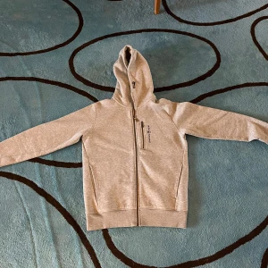 Grå hoodie från Sail Racing - Snygg ljusgrå hoodie från Sail Racing aldrig använd har testet 1-2 gånger. Den har hel dragkedja och broderad logga på bröstet. Tröjan har huva, två sidofickor och en bröstficka med dragkedja. Mjuk insida i fleece och klassisk passform. Perfekt för kyliga dagar.