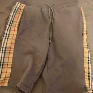 Grå Burberry shorts - Snygga grå shorts från Burberry med klassiska rutiga paneler i beige, svart och rött längs sidorna. Mjuk bomullskänsla, elastisk midja med snörning och avslappnad passform. Perfekta för en chill och trendig look.