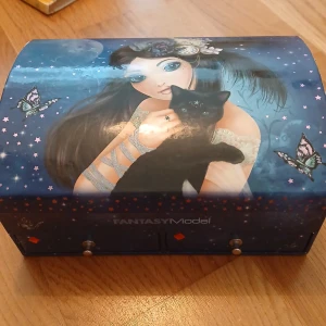 Fantasy Model smyckeskrin med spegel - Blått smyckeskrin med magiskt motiv av tjej, svart katt och fjärilar. Utsidan har stjärnor och nattblå bakgrund, insidan har spegel och flera fack med illustrationer av fåglar och enhörning. Perfekt för att förvara ringar, armband och halsband.