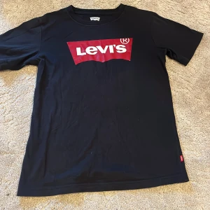 Svart Levi's t-shirt med röd logga - Svart t-shirt från Levi's med klassisk röd och vit logga framtill. T-shirten har rund hals, korta ärmar och är gjord i mjuk bomull. Perfekt för dig som gillar en enkel men ikonisk streetstil.