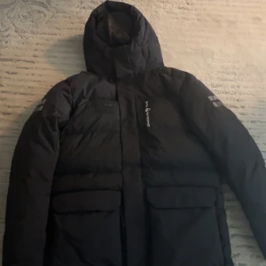 Svart dunjacka från Sail Racing L - Säljer en svart dunjacka från Sail Racing i storlek L. Jackan har huva, stora fickor framtill och logga på ärmen. Fylld med Allied Feather + Down, 500 fill power, vilket gör den riktigt varm och perfekt för kalla dagar. Snygg och stilren design med diskreta detaljer. Använt en gång!! Finns kvitto, en knapp har gått sönder, skriv privat för en mer tydlig bild. Annars syns det inte. 