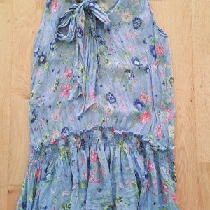 Blommig blå klänning från Zara - Supersöt blå kortklänning från Zara Woman med blommigt mönster i rosa, grönt och vitt. Klänningen är ärmlös, har v-ringning och en stor knytrosett vid halsen. Smockad midja och volangig nederdel ger en flowy känsla. Perfekt för sommarens alla tillfällen!