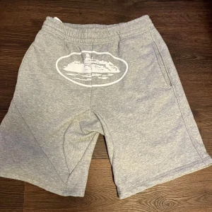 Grå sweatshorts från corteiz  - Grå sweatshorts från CORTEIZ med elastisk midja och snörning. Mjuk bomullsmix, normal passform och fickor på sidorna samt en bakficka. Snyggt vitt tryck på ena benet. Perfekta för chill eller träning.
