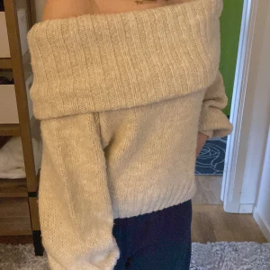 Beige offshoulder stickad tröja - Supermysig beige stickad tröja med bred ribbad kant som sitter offshoulder. Tröjan har långärmade ärmar och en loose fit, perfekt för dig som gillar en chill och trendig look. Passar perfekt till jeans eller kjol för en snygg outfit.