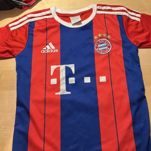 Bayern München Lewandowski tröja - Klassisk Bayern München fotbollströja från Adidas med Lewandowski och nummer 9 på ryggen. Tröjan har breda röda och blå vertikala ränder, vita detaljer och klubbmärke på bröstet. Korta ärmar och sportig passform, perfekt för fans av tysk fotboll.