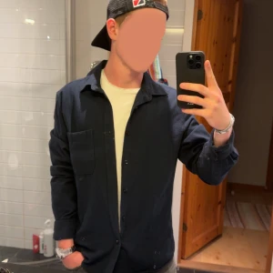 Mörkblå overshirt med bröstficka - Snygg mörkblå overshirt med klassisk krage och stora knappar framtill. Skjortan har en bröstficka på vänster sida och är tillverkad i ett mjukt tyg som passar perfekt över en t-shirt. Enkel och stilren design som funkar till många outfits.