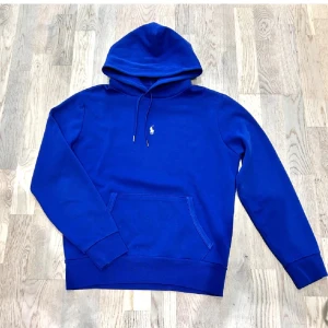 Blå Polo Ralph Lauren hoodie - Snygg blå hoodie från Polo Ralph Lauren med klassisk vit logga på bröstet. Tröjan har huva med snörning, känguruficka och ribbade muddar. Perfekt för dig som gillar stilrena och bekväma plagg med premiumkänsla.