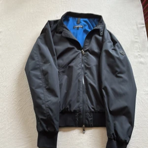 Mörkblå bomberjacka från J.Lindeberg - Snygg mörkblå bomberjacka från J.Lindeberg med blått innerfoder. Jackan har dragkedja framtill, ribbade muddar vid ärmslut och midja samt klassisk krage. Perfekt för dig som vill ha en stilren och sportig look. Orginal pris 3000kr. Använd 1 gång.